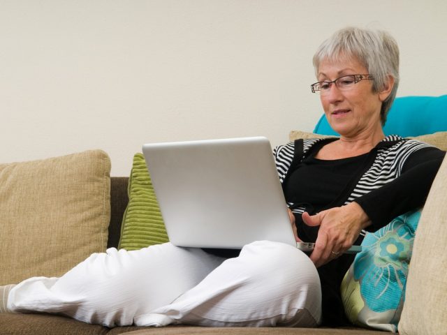 Senior Woman Working On A Laptop, Relaxed On The Couch